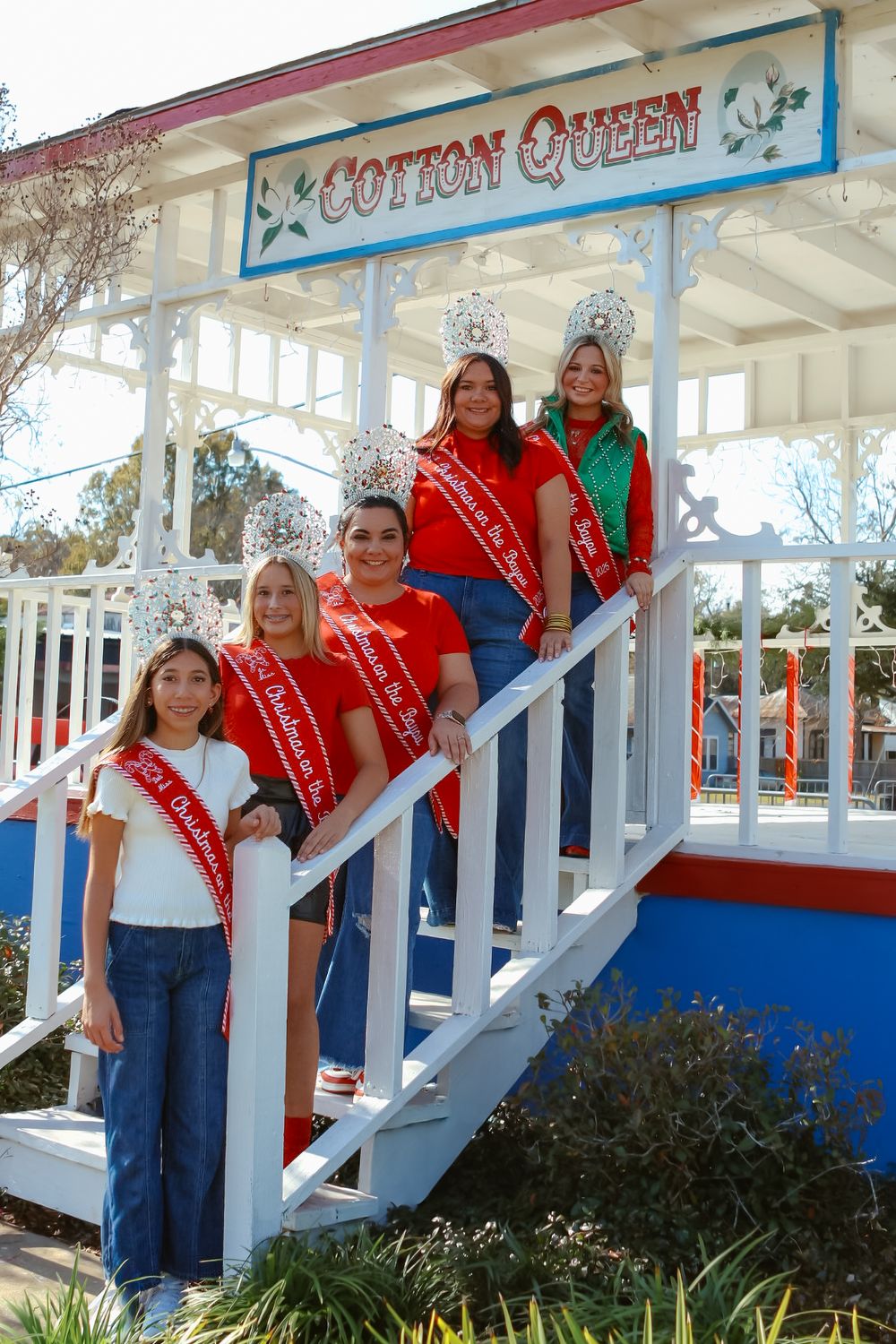 Group wearing red sashes and crowns pose together on a white porch with blue accents.