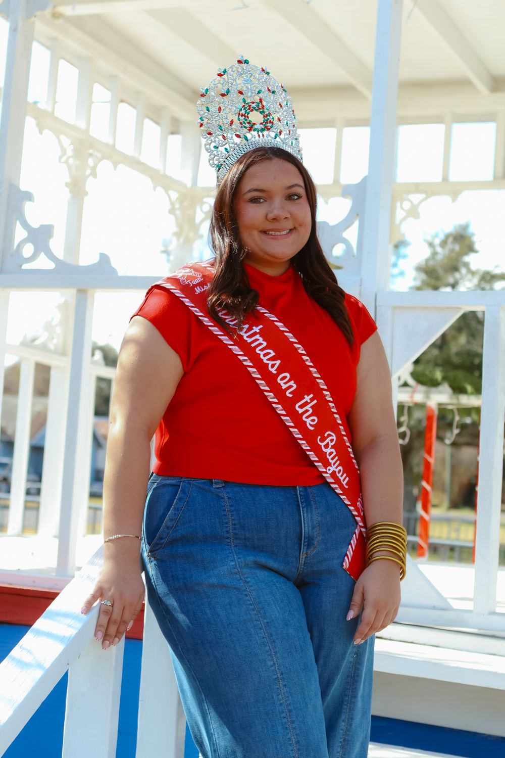 Person in red shirt and jeans poses on white porch with decorative crown.