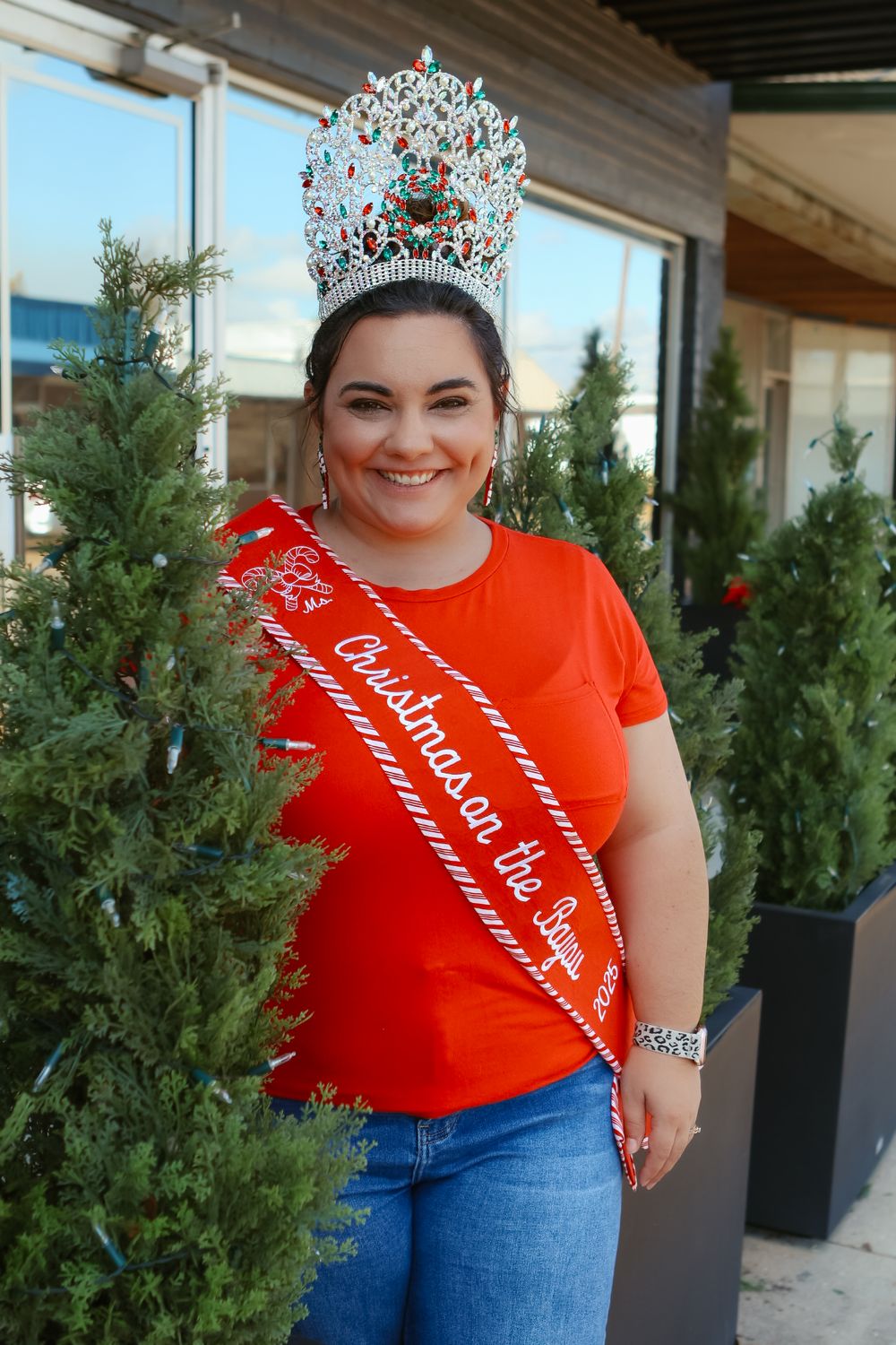 Person in red shirt and jeans poses by potted evergreen trees wearing crown and sash.