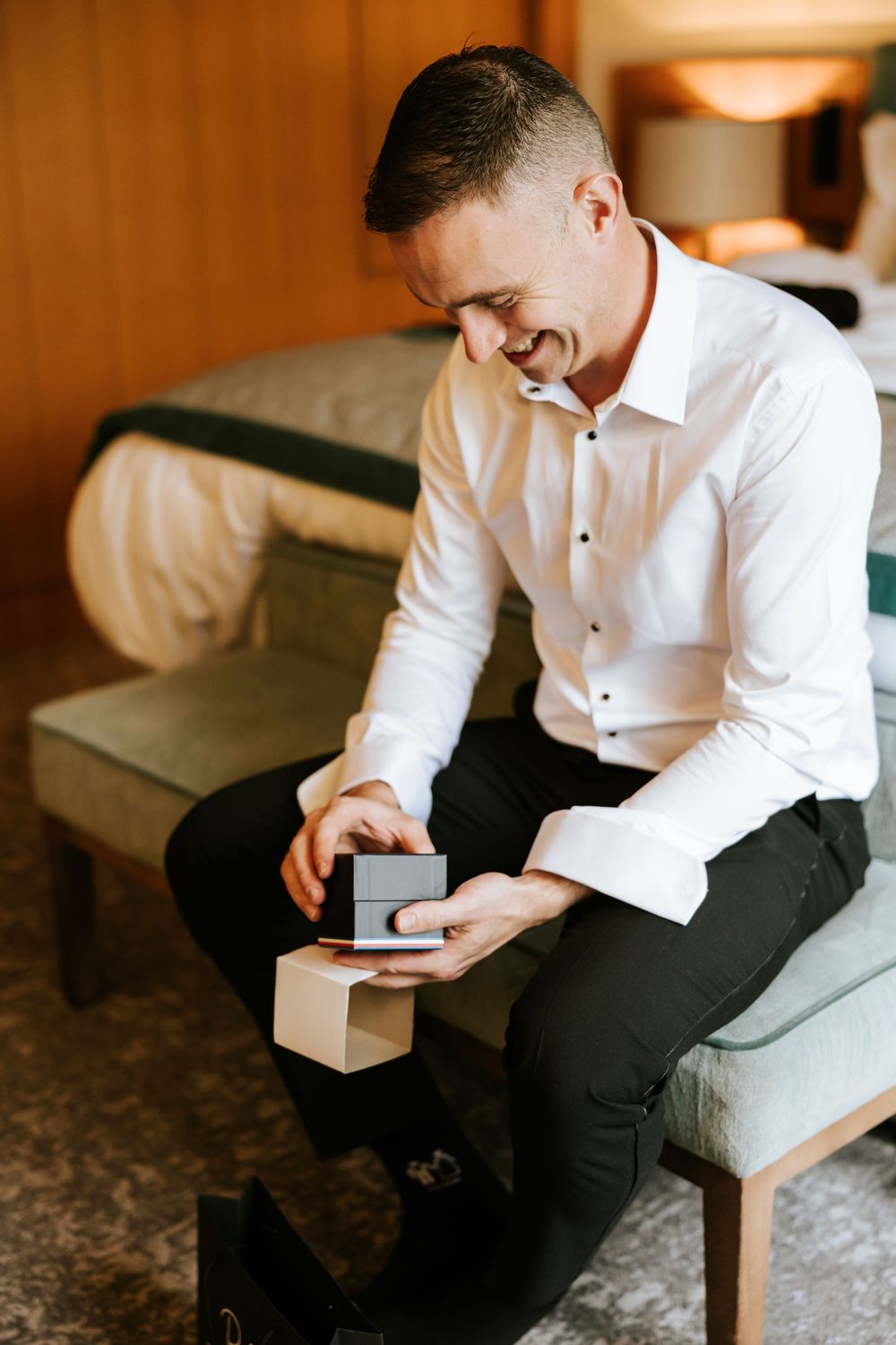 Someone in a white dress shirt and black pants sits on a couch while opening a small gift box. Farnham estate