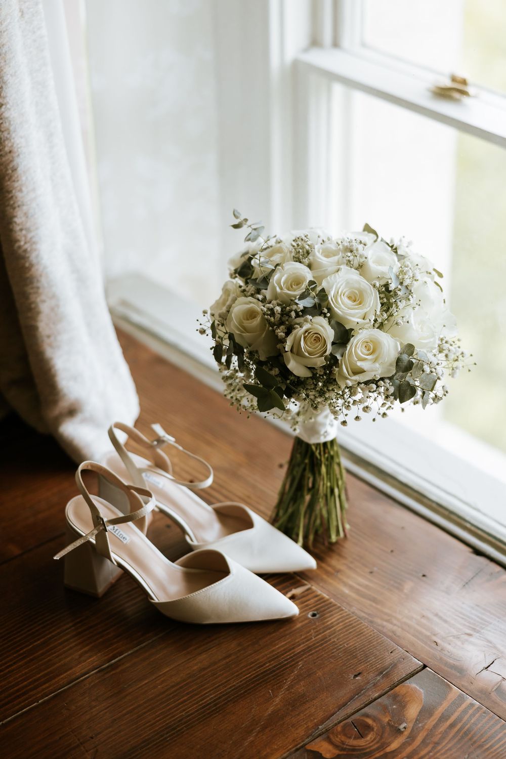 White bridal heels and a classic white rose bouquet with baby's breath sit on a wooden windowsill in natural light. Farnham estate