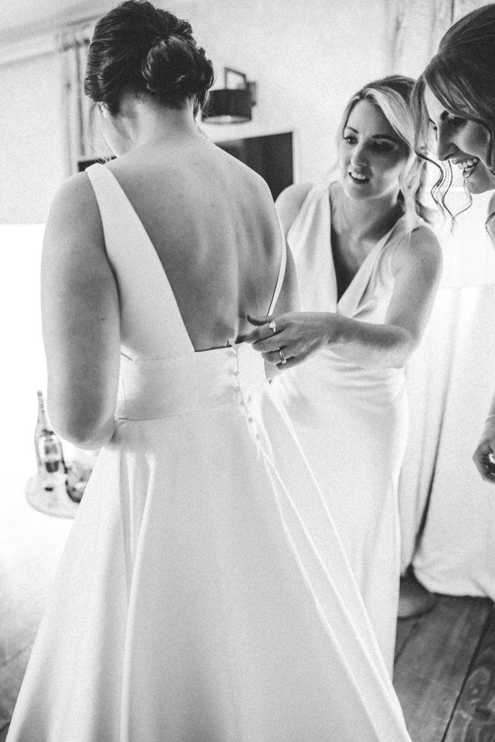 Black and white photo showing final wedding dress preparations with bridesmaids helping adjust the gown's back details. Farnham estate