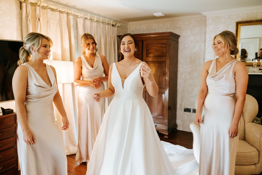 A joyful wedding preparation scene with bridesmaids in blush dresses surrounding a bride in a classic white gown. Farnham estate
