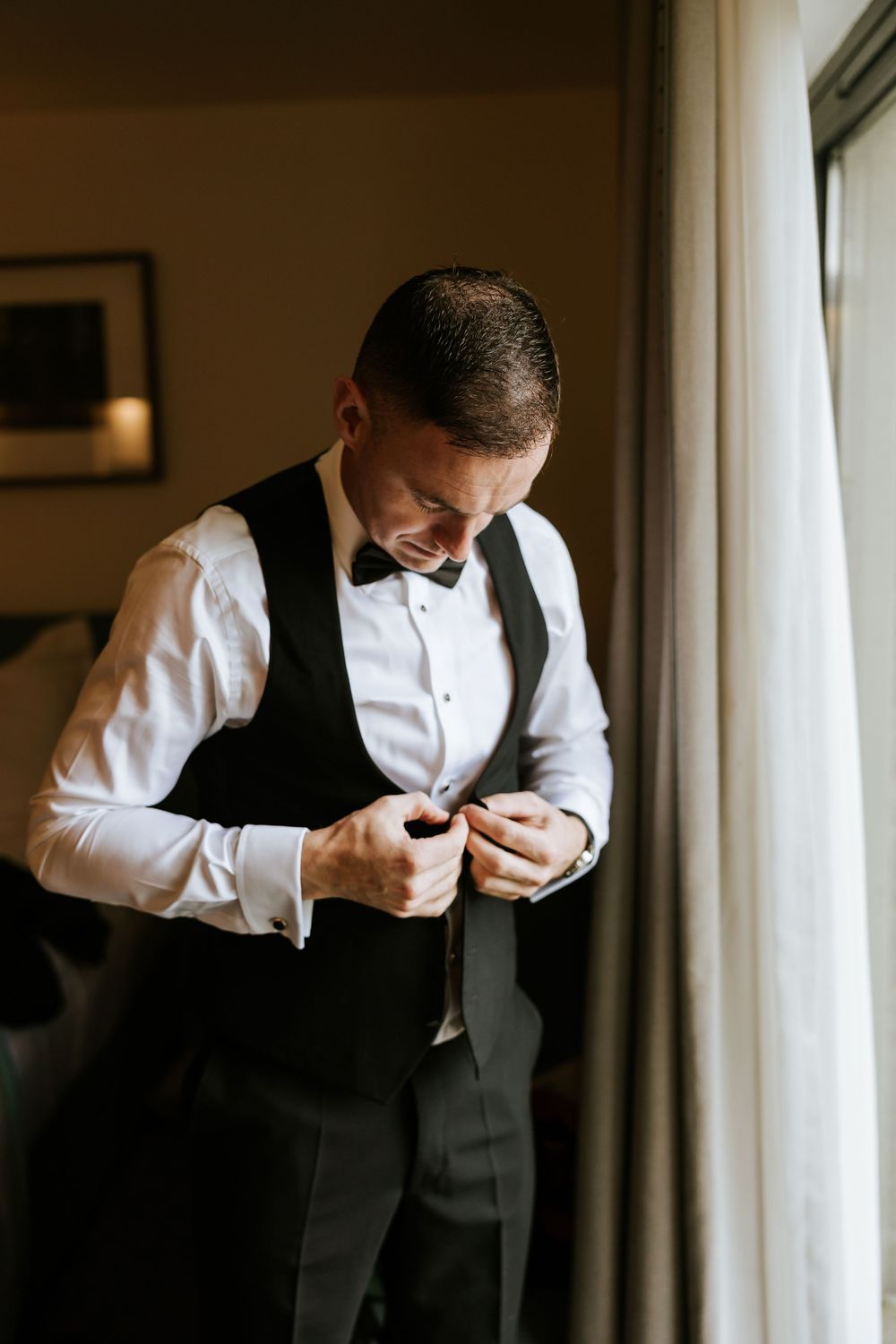 Close-up shots of a groom getting dressed in formal attire by a window in warm lighting. Farnham estate