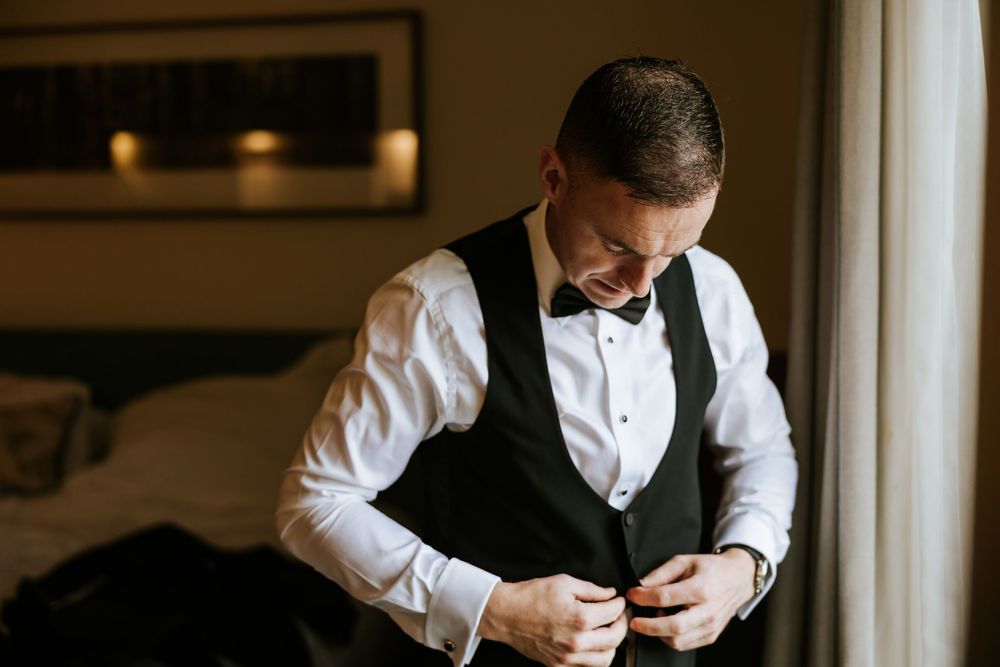 Groom in white dress shirt and black vest adjusts his buttons while getting ready by window light. Farnham estate