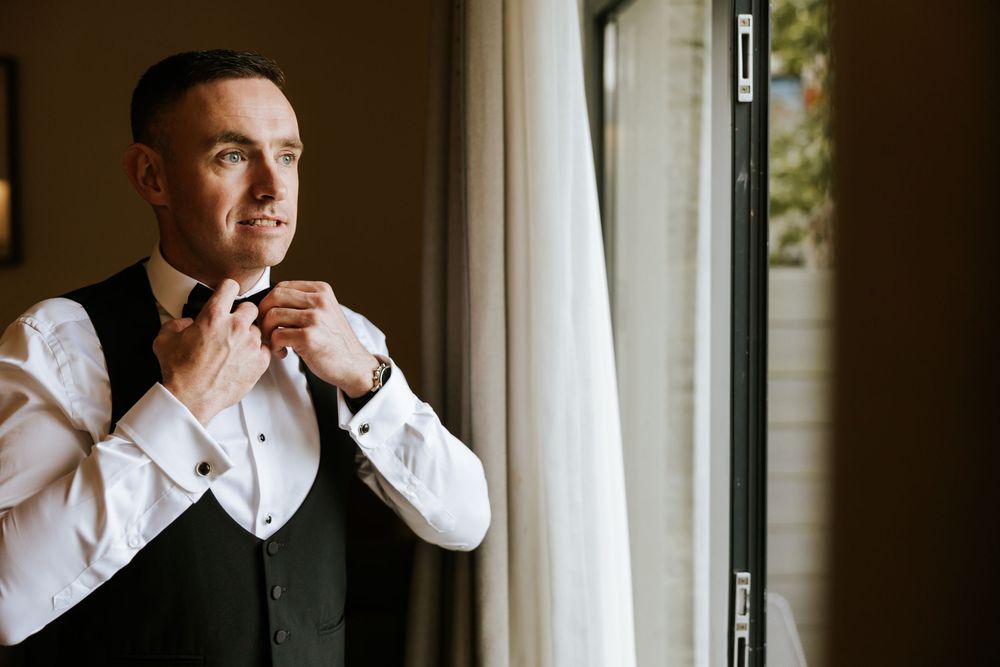 A person in a formal vest and white shirt adjusts their cuff while standing by a window in natural light. Farnham estate