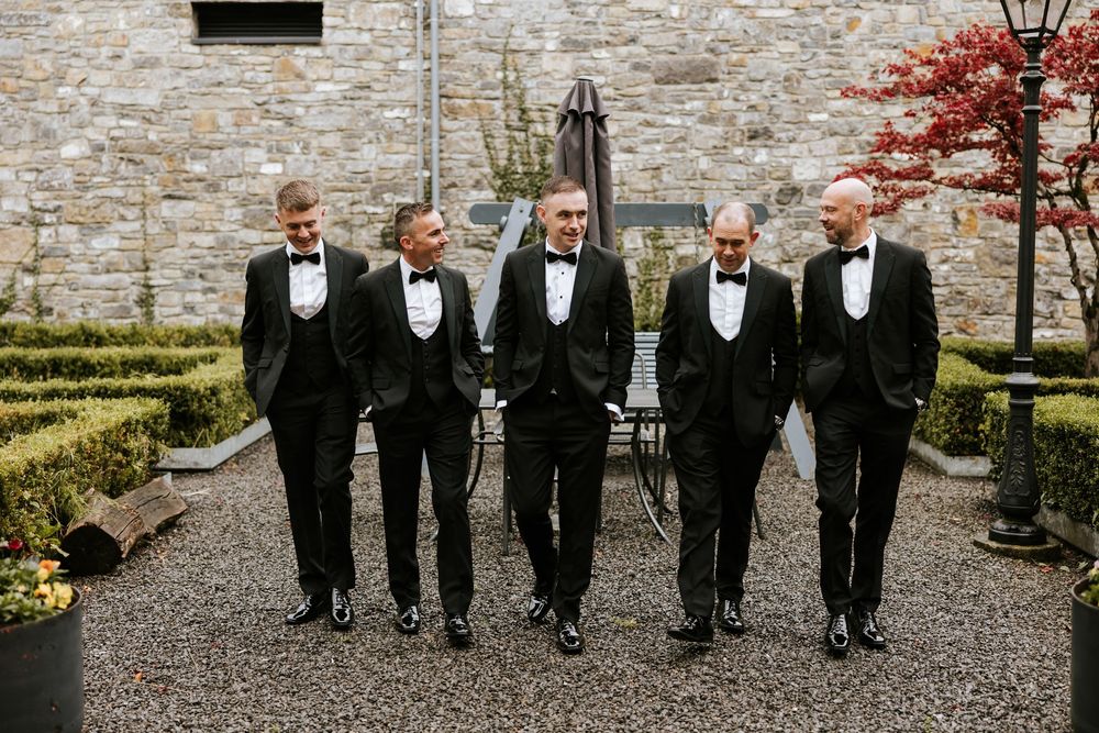 Groomsmen in black tuxedos walking together on a gravel path near a stone building and autumn trees. Farnham estate