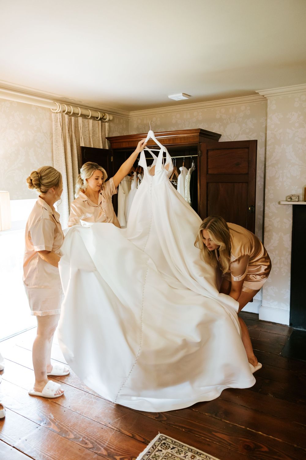 A group helps with a flowing white wedding dress in a room with hardwood floors and vintage furniture. Farnham estate