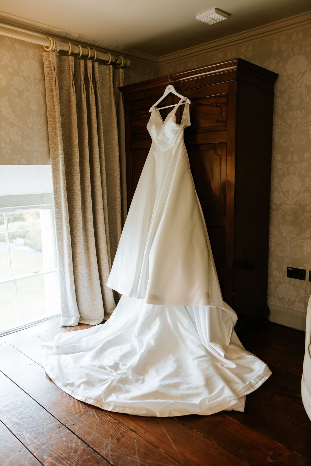 A white wedding dress hangs elegantly in a room with wooden floors and cream colored curtains. Farnham estate