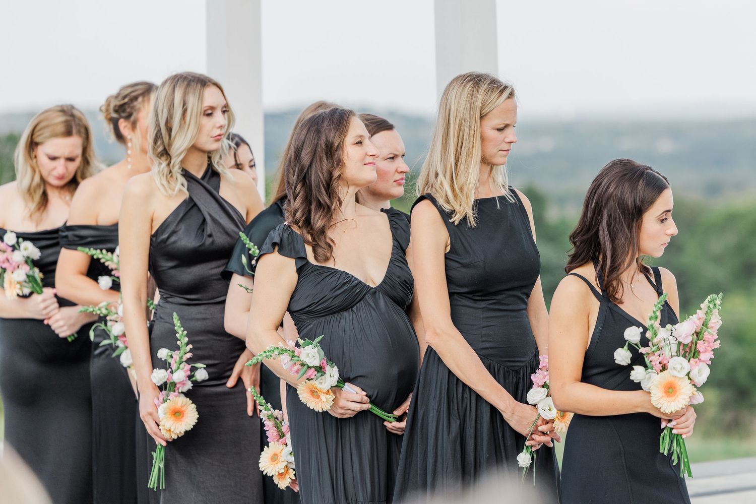 Bridesmaids in elegant black dresses holding bouquets stand in a row during an outdoor wedding ceremony.