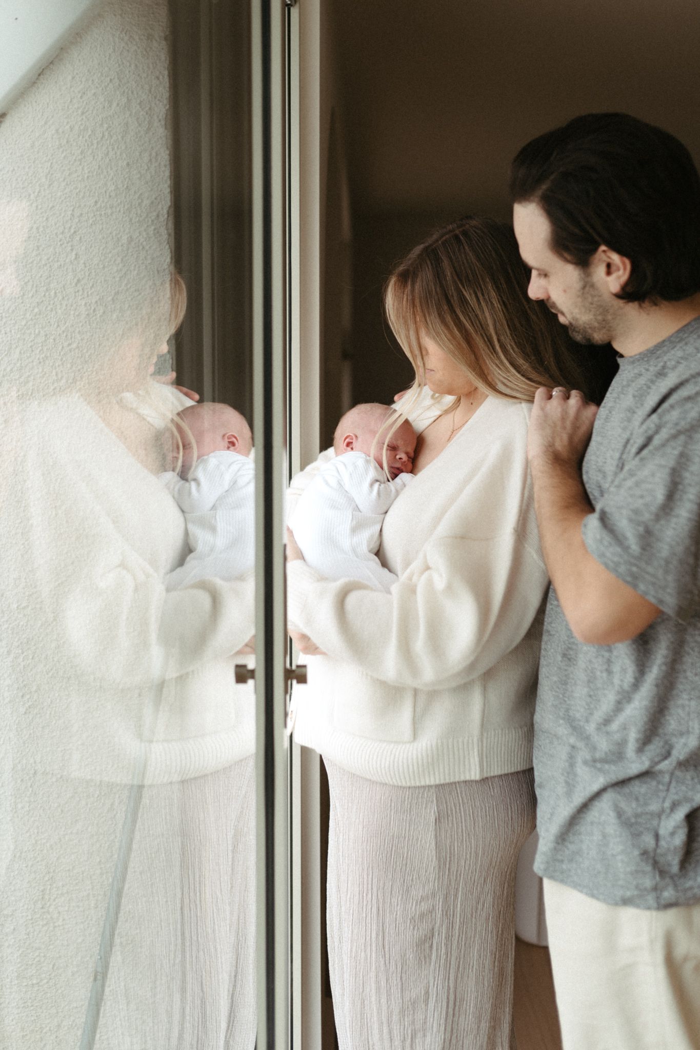 A couple stands by a window with their newborn baby, creating a tender family moment in soft natural light.