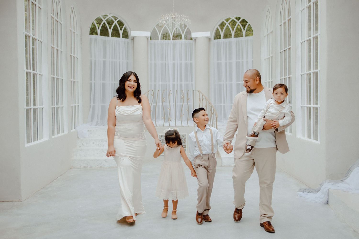 Family of five wearing matching white outfits pose together in an elegant white room with arched windows.