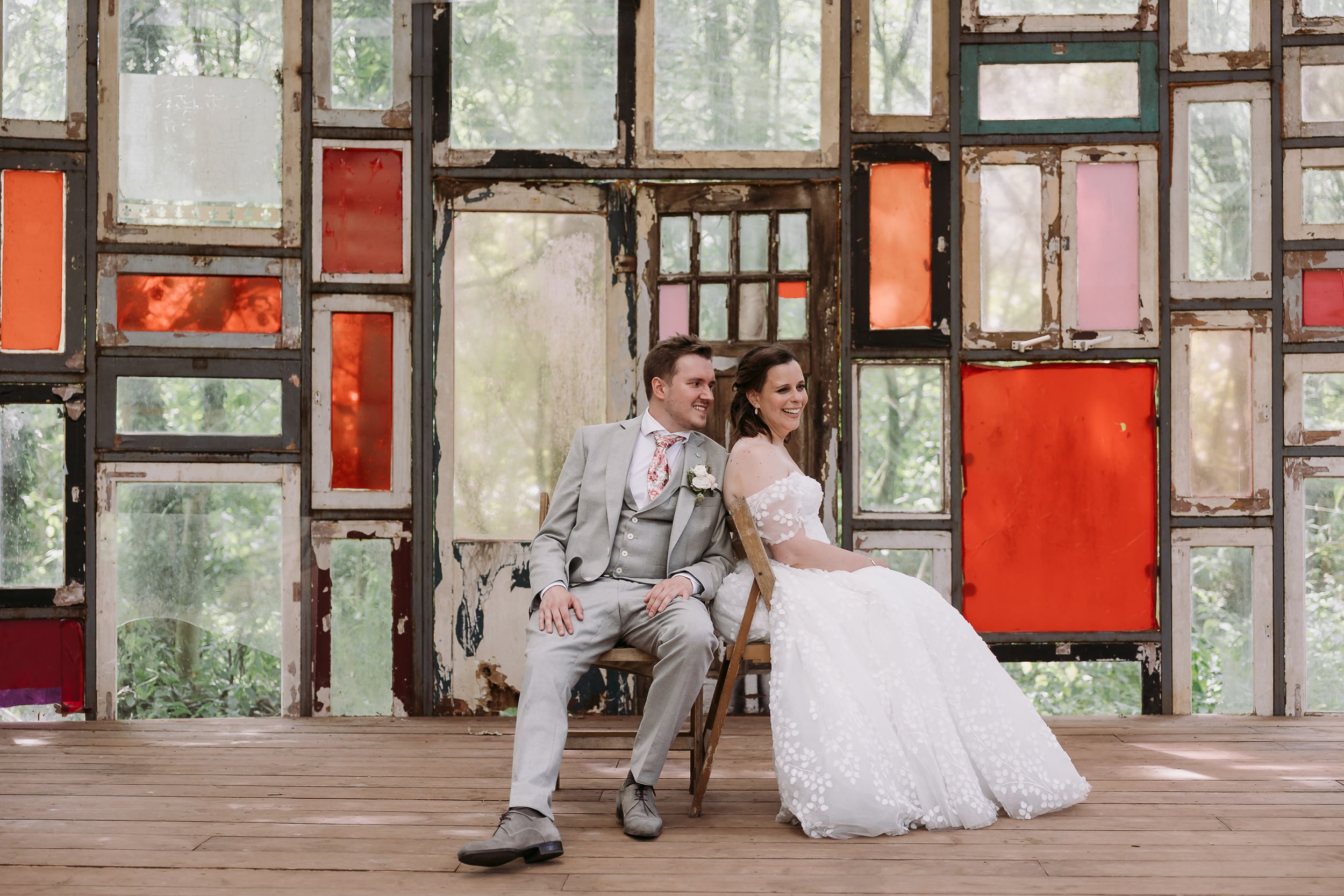 A couple in wedding attire sits together on a bench against a colorful geometric wall with glass panels.