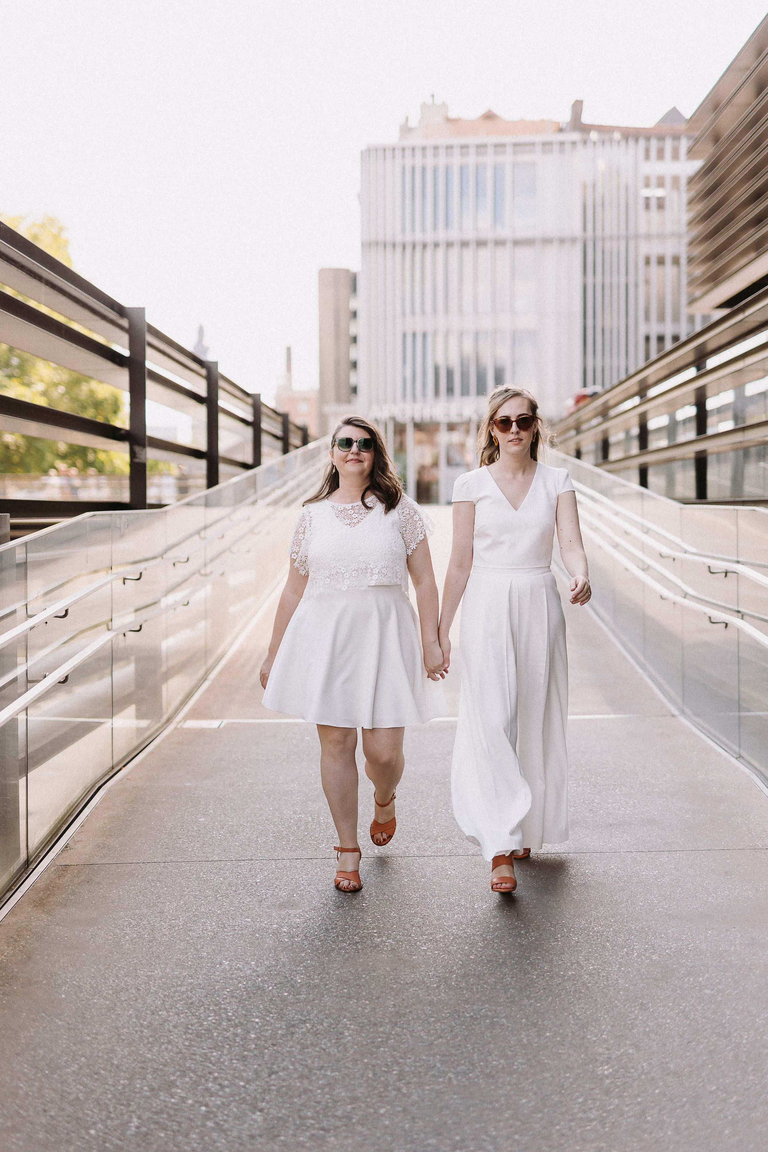 Two people in white dresses hold hands while walking down a modern urban pathway with metal railings.