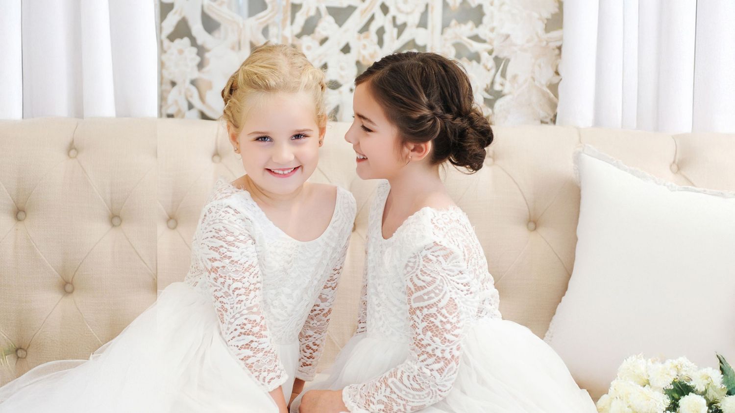 Two young flower girls in white lace dresses sit together smiling on a tufted cream sofa.