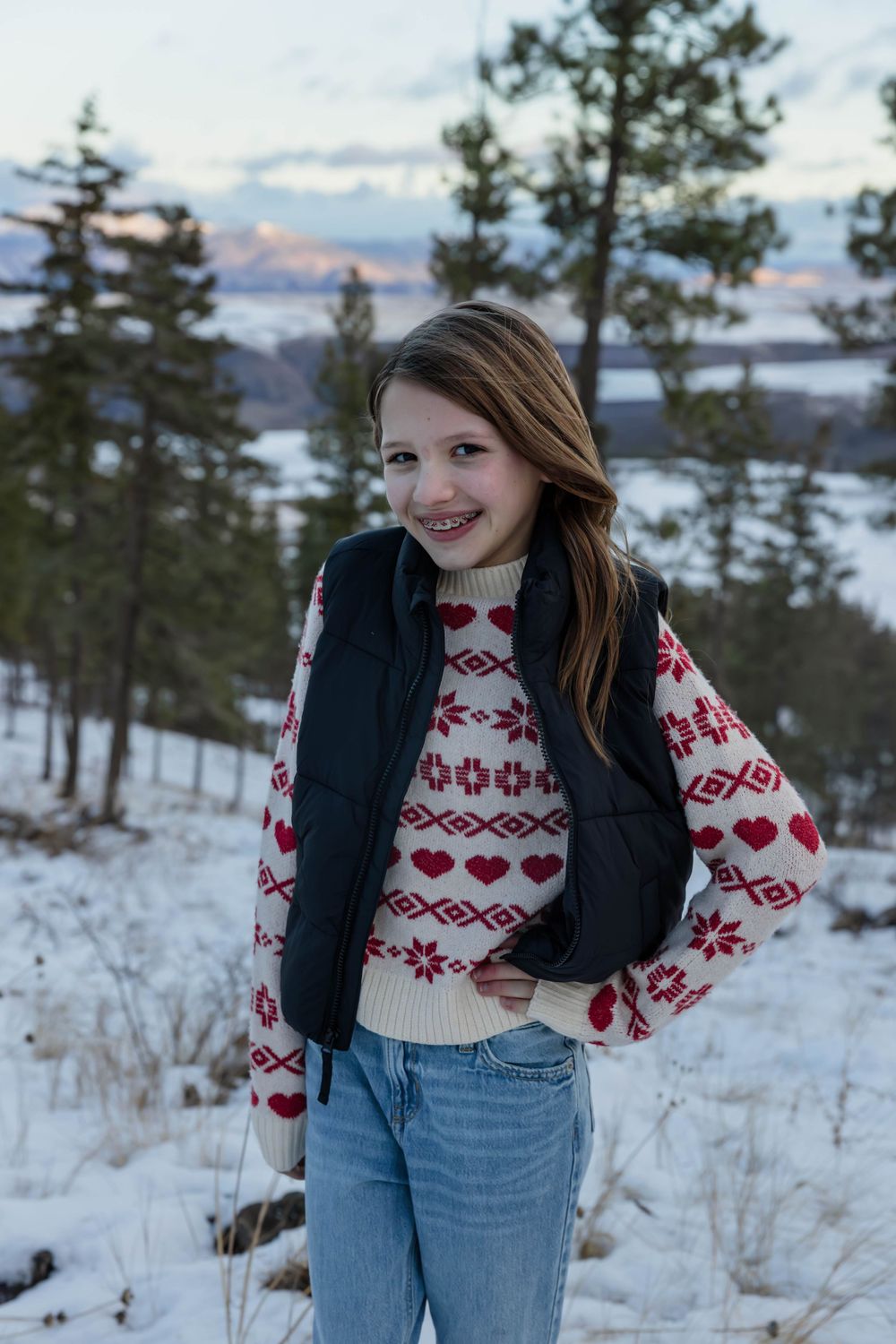 Person in a festive heart-patterned sweater and vest poses in a snowy mountain setting.
