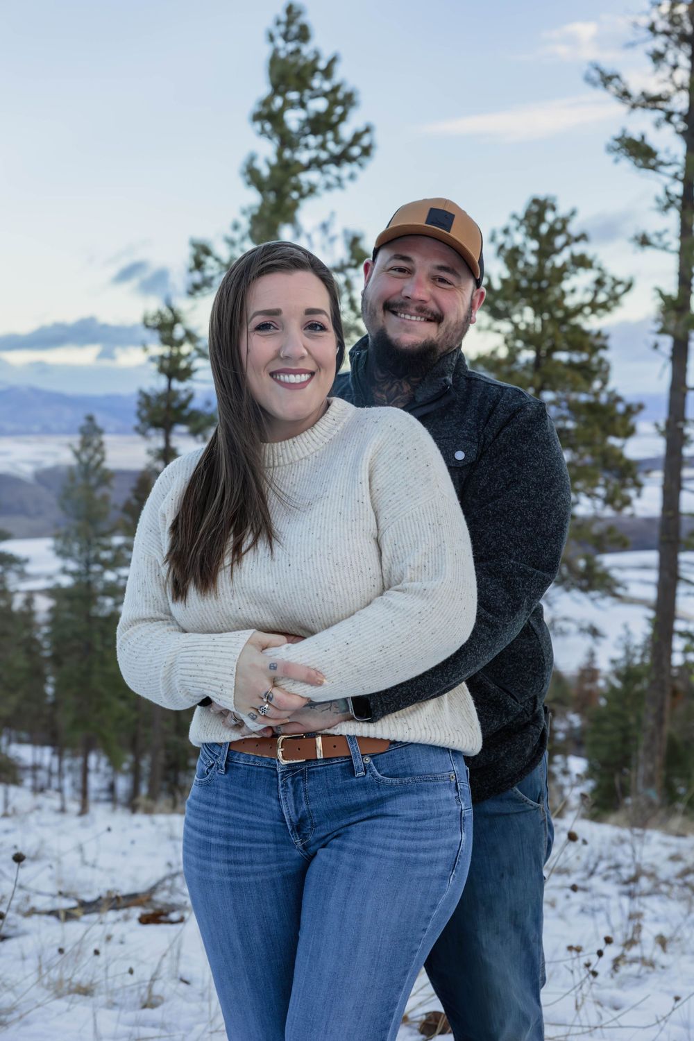 Couple embraces during a winter photoshoot with snow-covered mountains in the background.