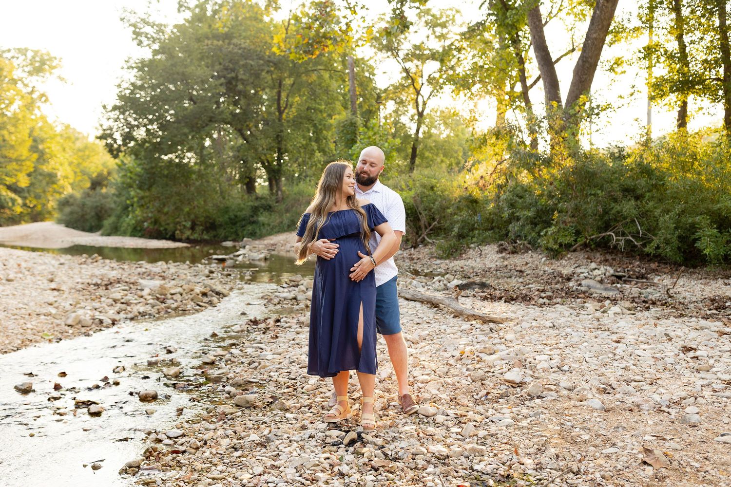A couple stands together on a rocky riverbank surrounded by autumn trees during a photo session.