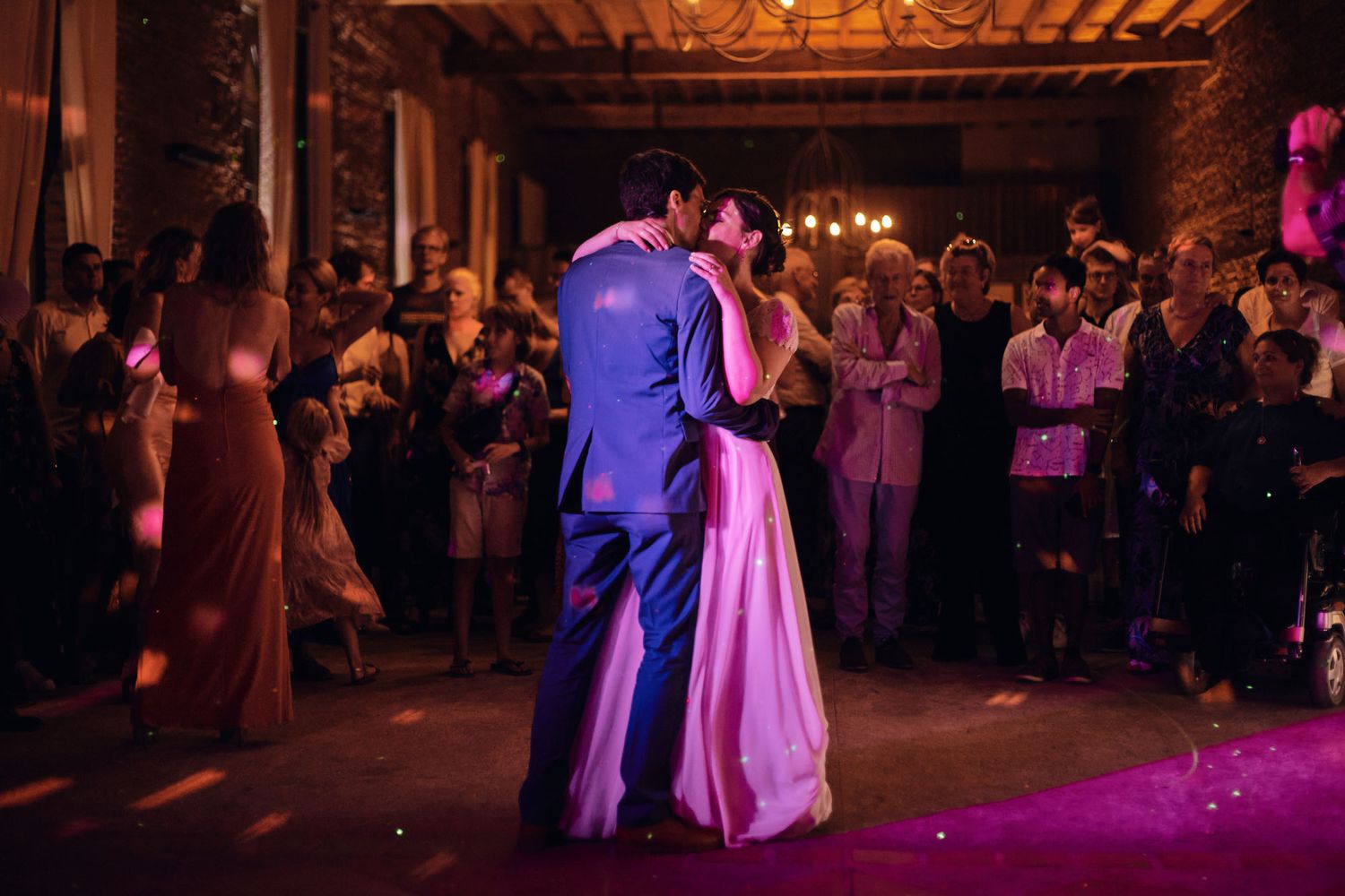 A couple shares their first dance surrounded by guests in a dimly lit reception venue.