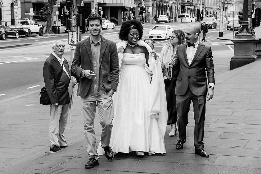 A wedding party walks down a city street in black and white, with the bride wearing a flowing white dress.
