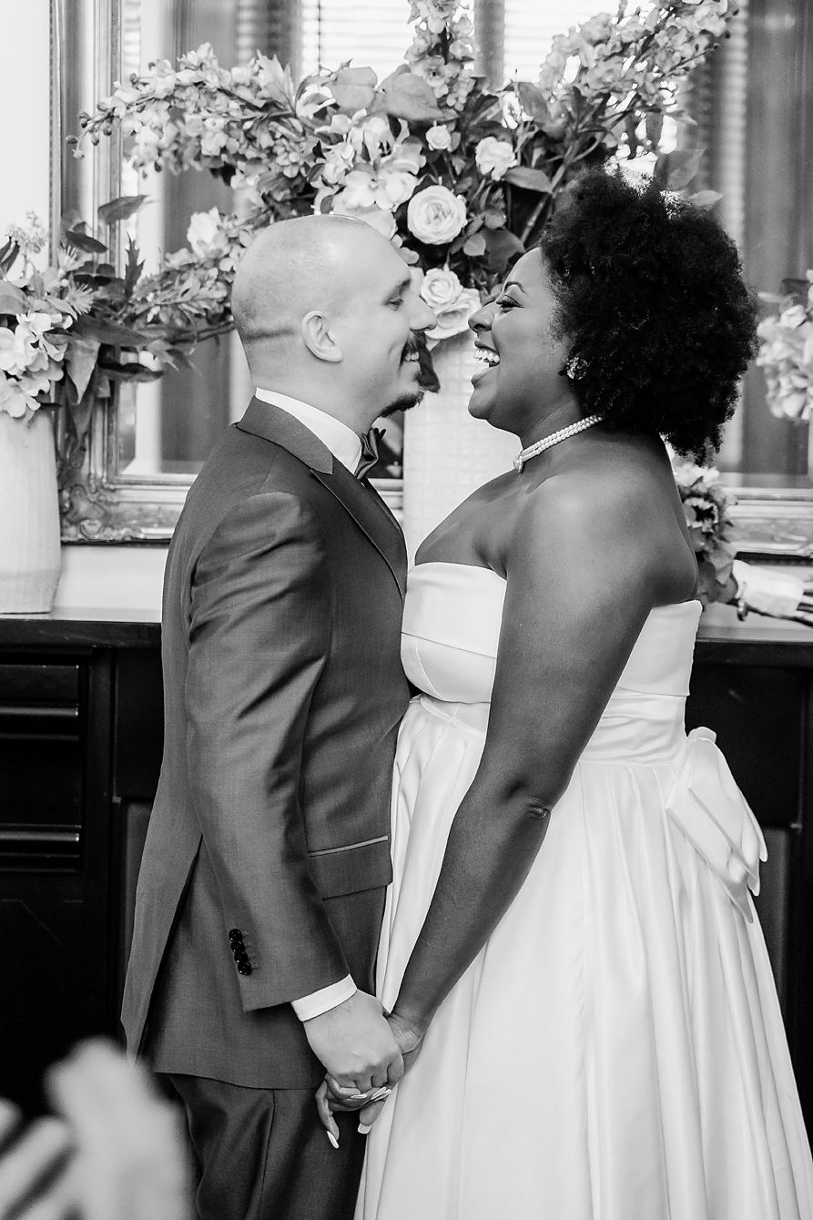 A black and white wedding photo showing a tender moment between newlyweds with floral arrangements in the background.