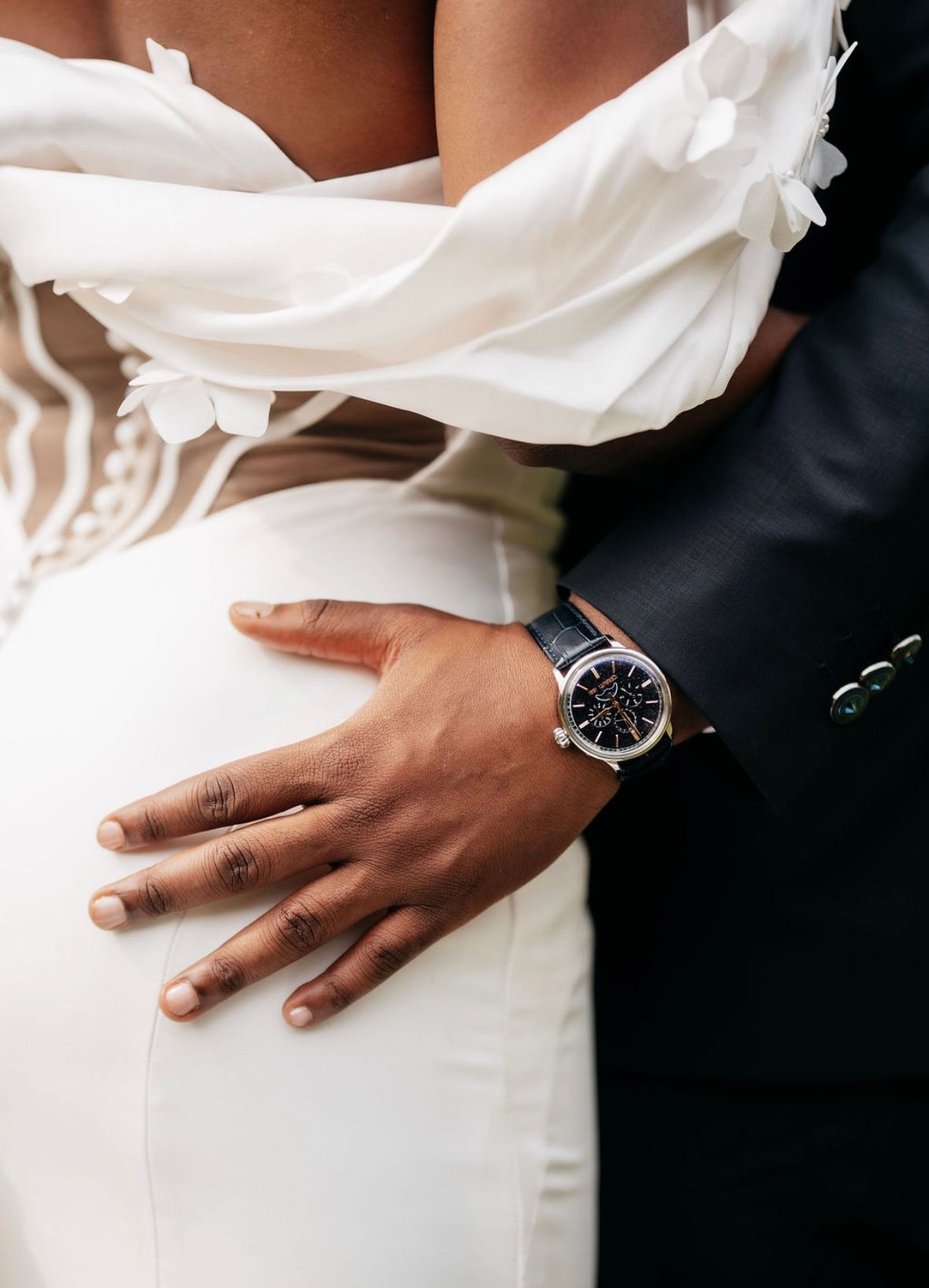 A close-up view of hands wearing wedding rings against a white dress and dark suit.