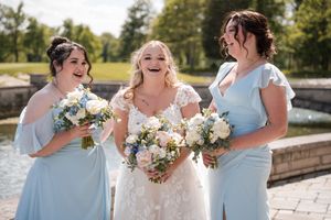 Three people in light blue dresses and white hold bouquets while laughing together during outdoor wedding celebration.