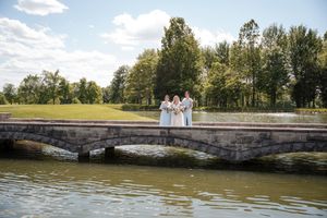 Three figures in white attire stand on a stone bridge overlooking a tranquil lake on a sunny summer day.