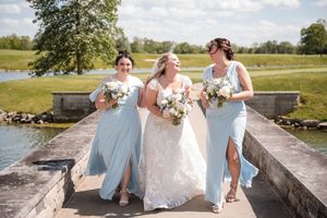 Three friends in light blue and white dresses walk together holding bouquets on a sunny day at an outdoor venue.