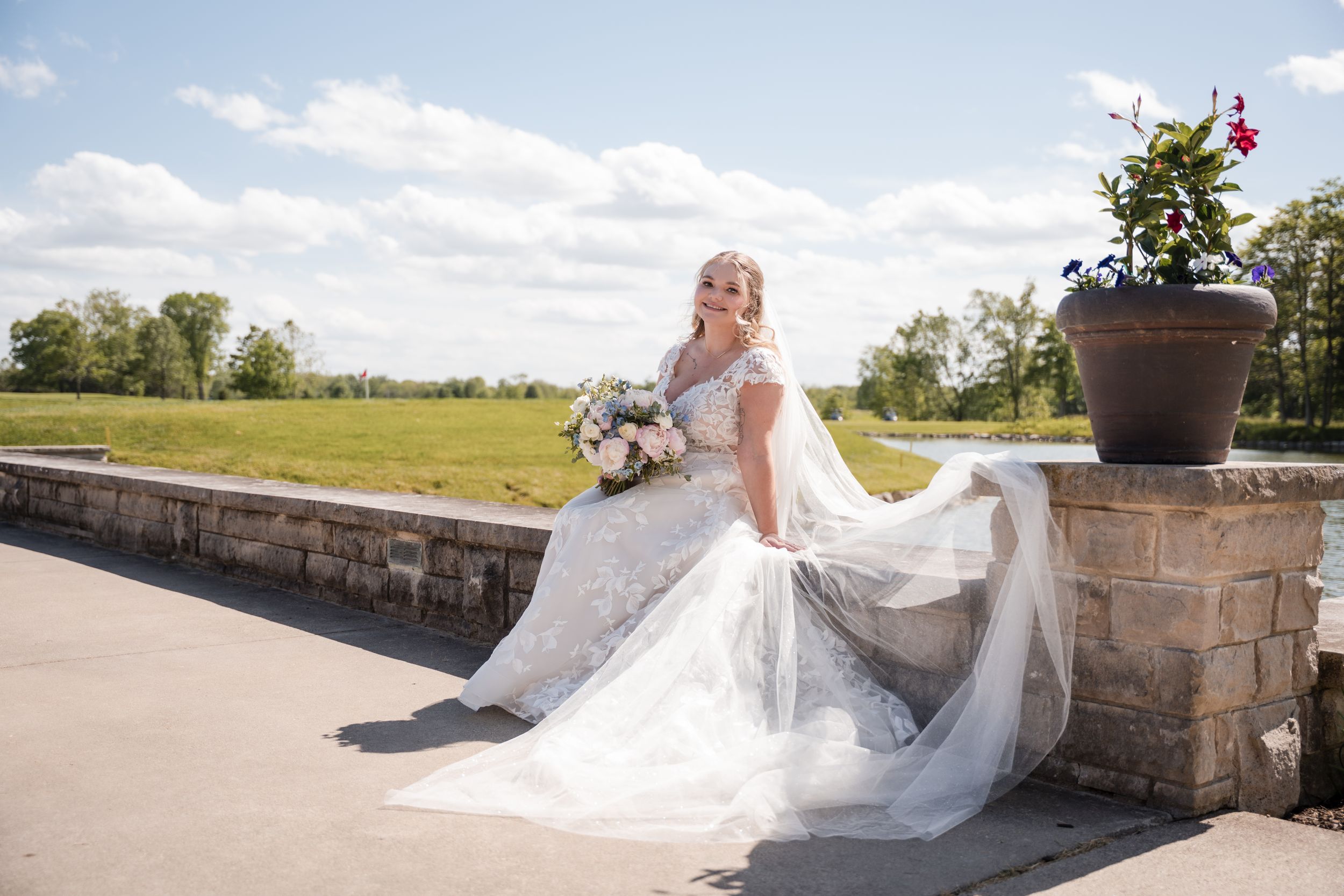 A white wedding dress flows elegantly across stone steps overlooking a sunny countryside landscape on a beautiful summer day.