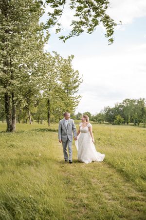 A newlywed couple walks together through a sun-lit meadow with trees in the background.