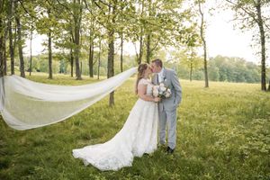 Two people in wedding attire embrace in a sunny field while a long veil flows in the breeze.