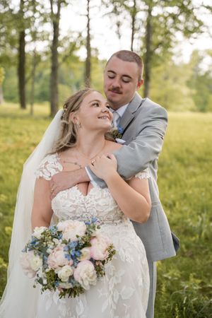 A romantic outdoor wedding photo in a sun-dappled field featuring a lace dress and beautiful pale bouquet with blue accents.