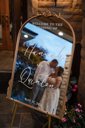 An elegant mirrored welcome sign displays wedding details with decorative gold frame and floral accents.