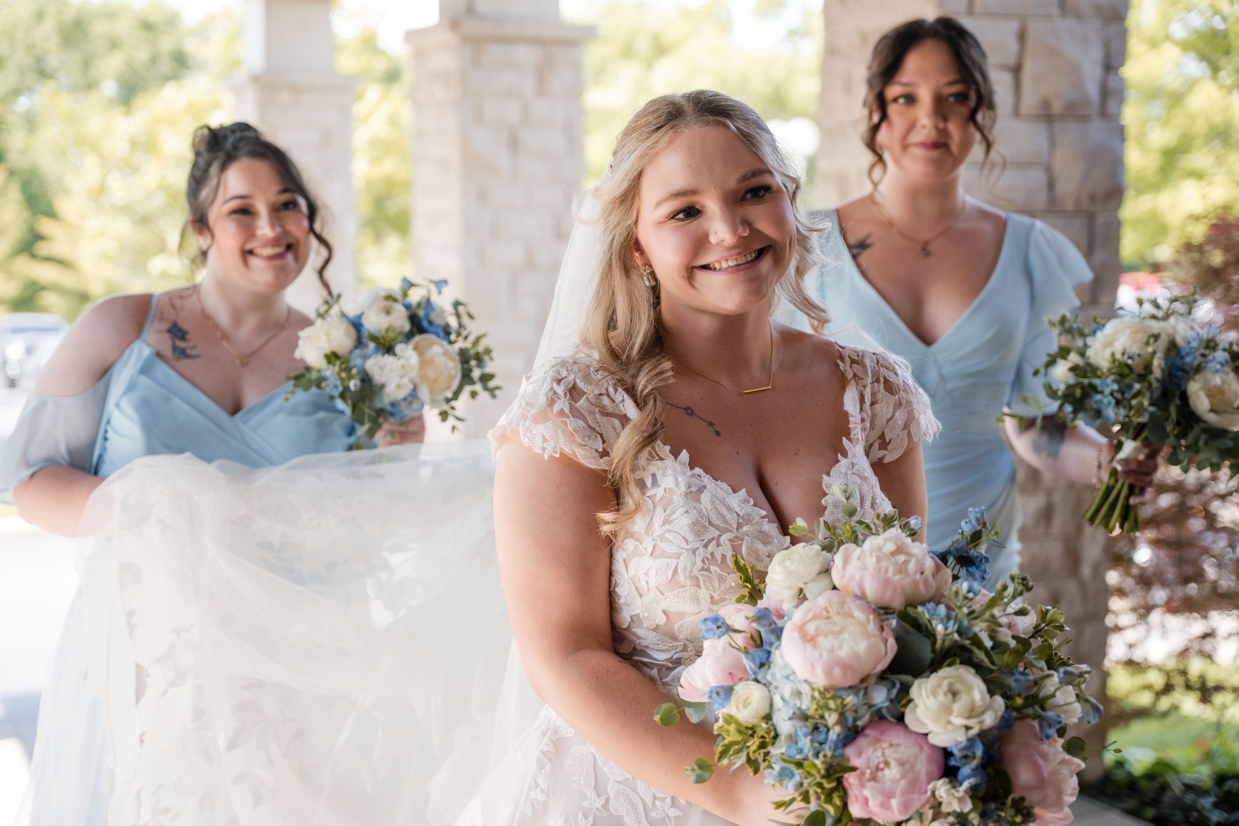 Three women in blue and white dresses hold beautiful pastel floral bouquets while smiling outdoors.