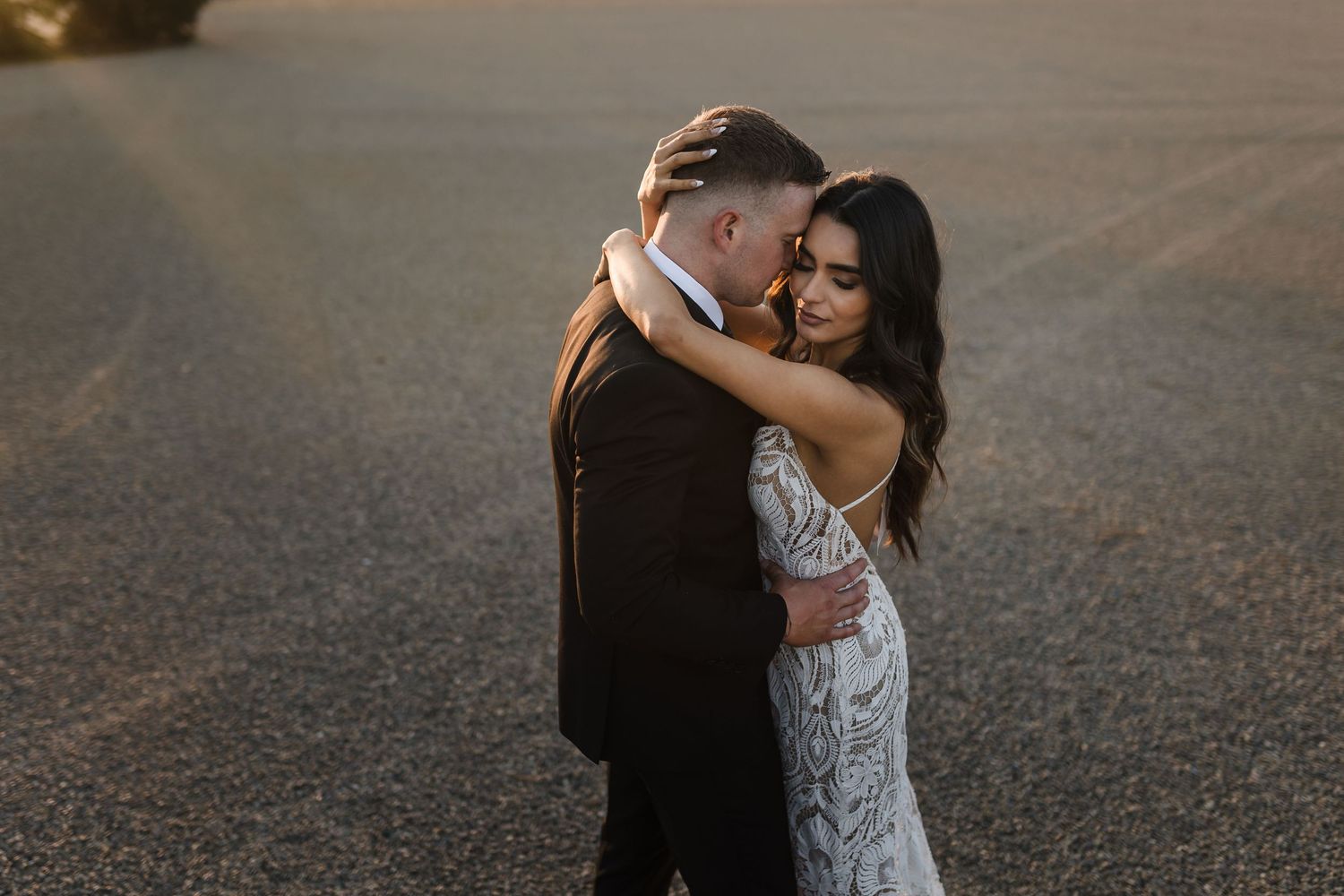 A romantic couple embraces on a paved surface during golden hour, the bride wearing a delicate lace wedding dress.