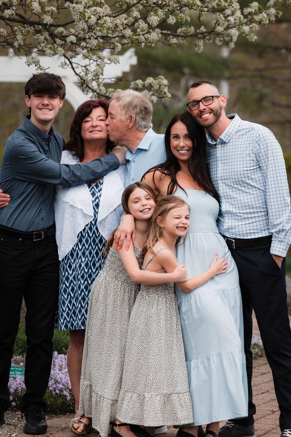 A multigenerational family poses together under blooming spring tree branches with two young girls in dresses.