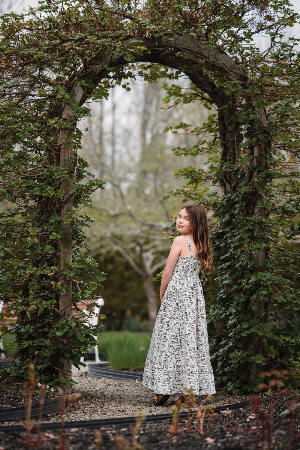 A figure in a flowing gray dress stands beneath a lush garden archway on a gravel path.