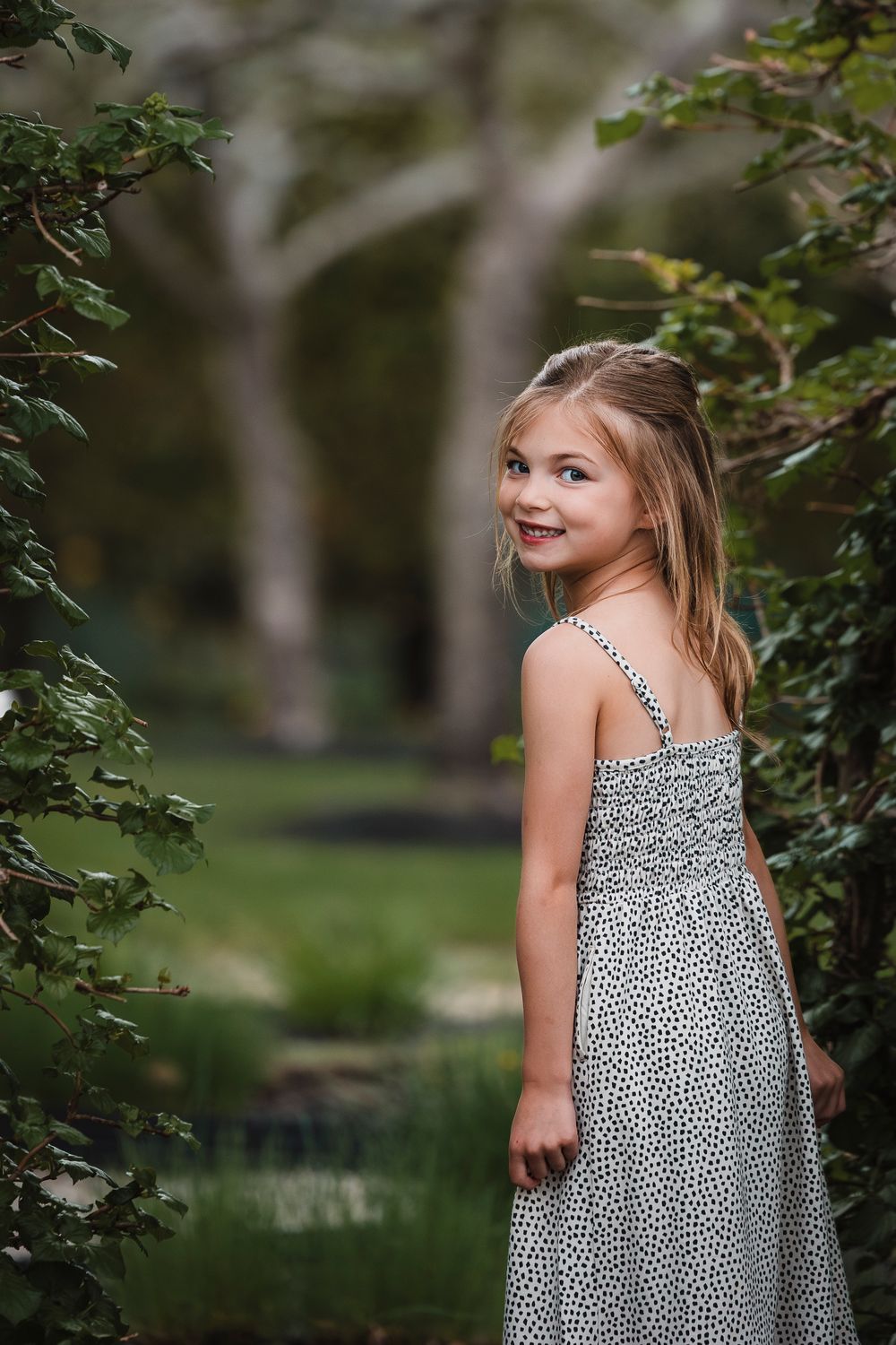 A young girl in a polka dot dress poses in a garden setting with a genuine smile.