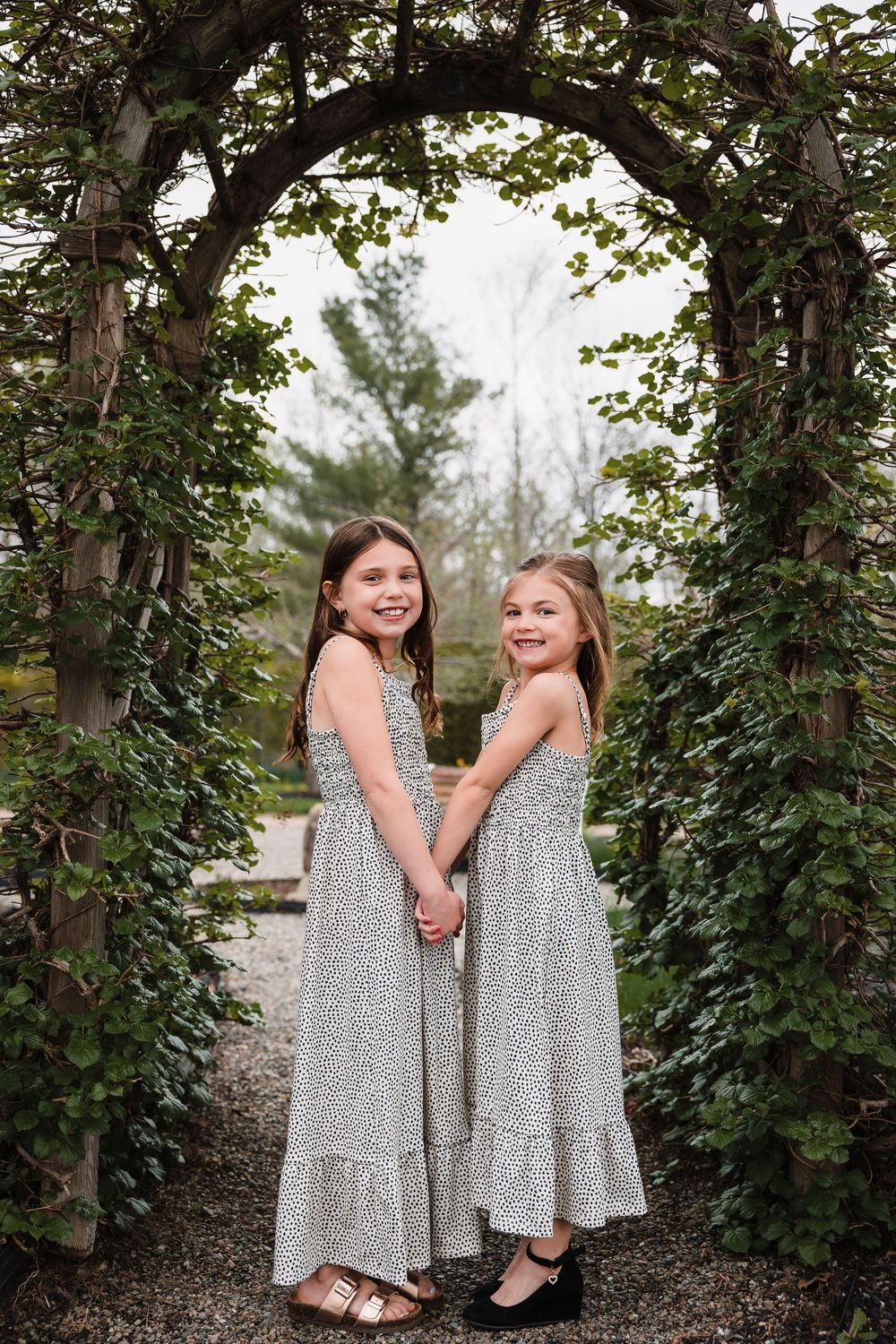 Two children in matching light floral dresses stand holding hands under a garden arch with green vines.