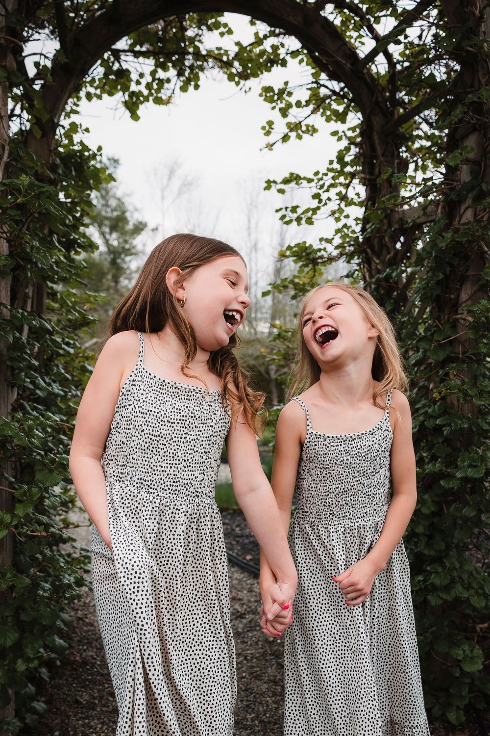 Two friends in matching patterned dresses share a joyful laugh while holding hands under a green archway.