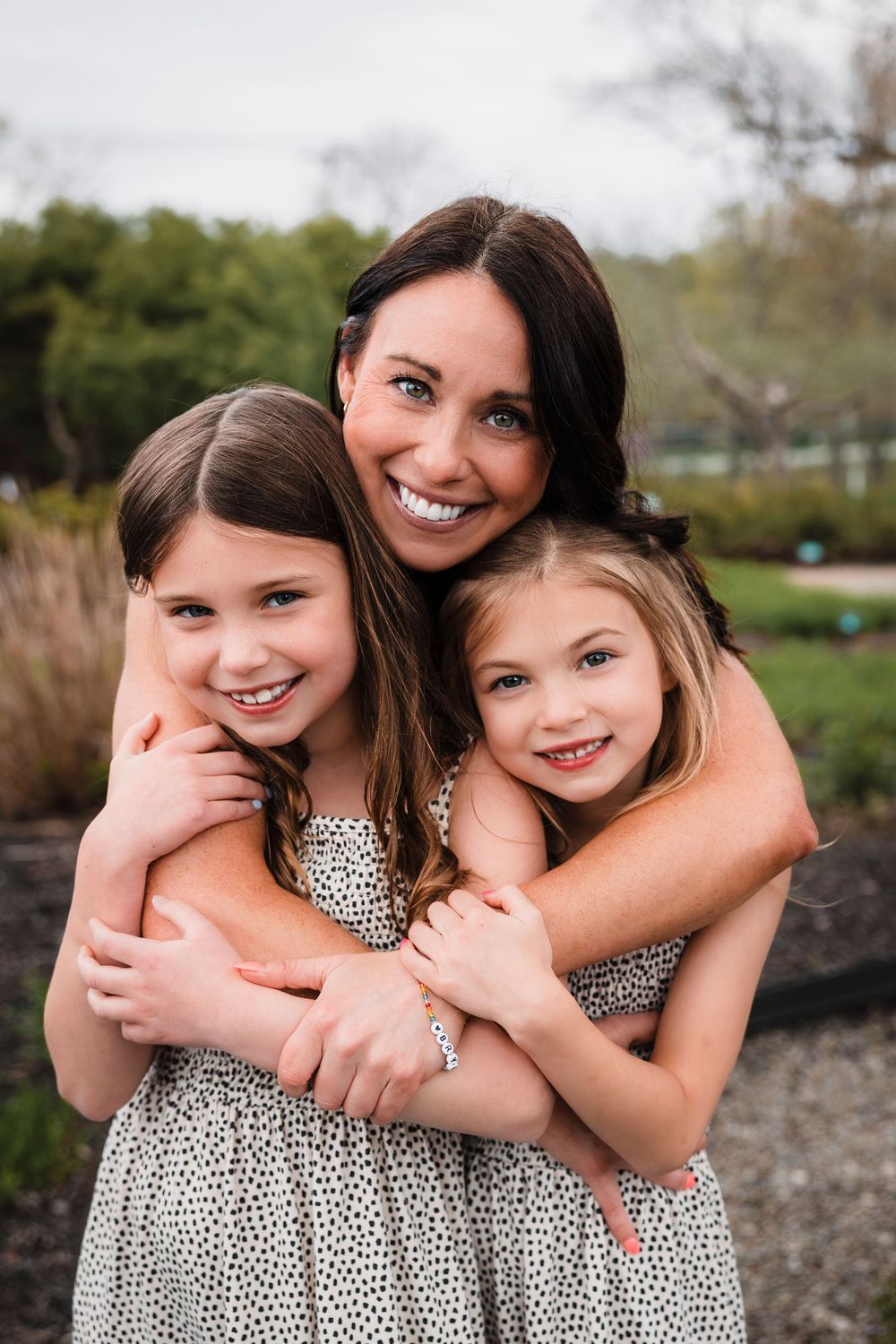 Three people embracing and smiling joyfully while wearing matching white and black polka dot dresses outdoors.