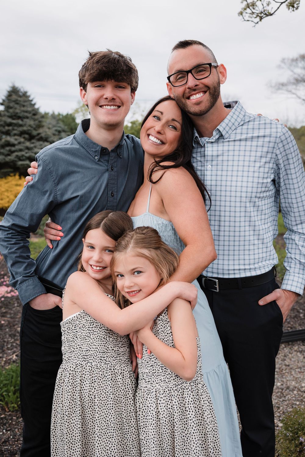 Family of five posing outdoors in coordinated outfits with pine trees and autumn foliage in background.