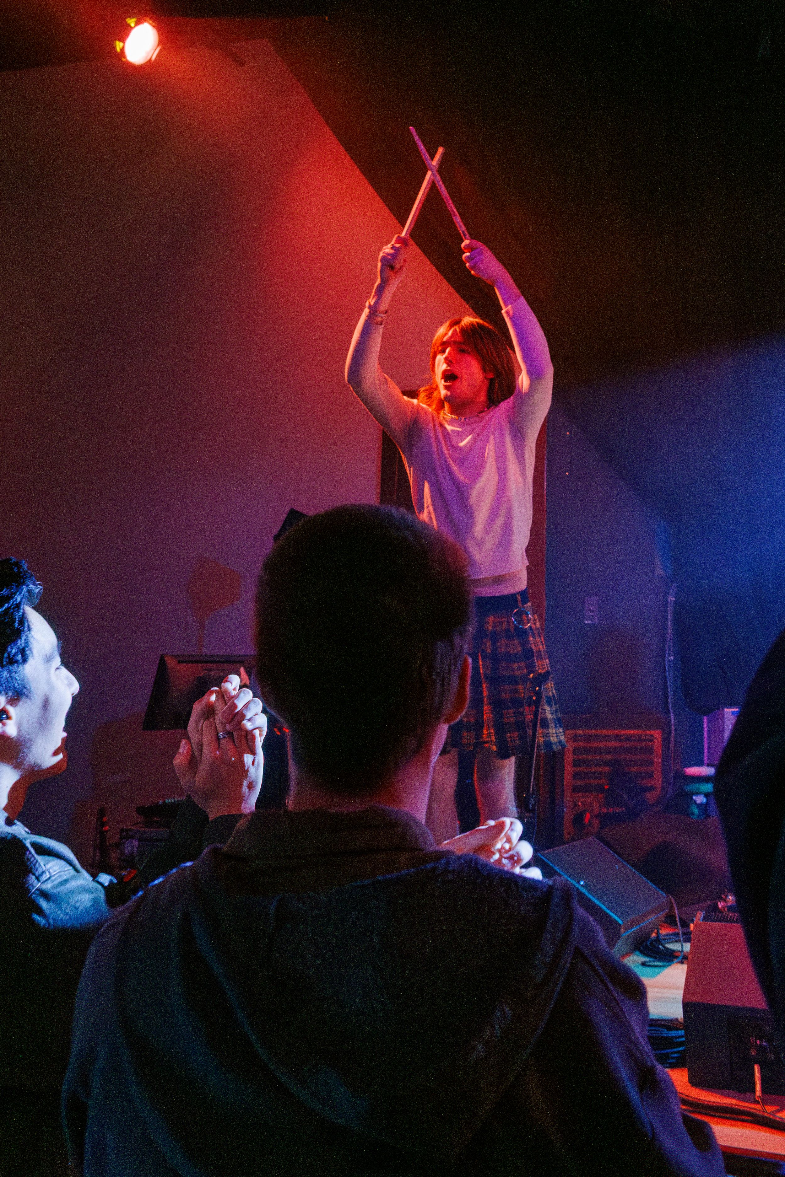 A performer in a white t-shirt holds drumsticks up while standing on stage under dramatic red and blue lighting.