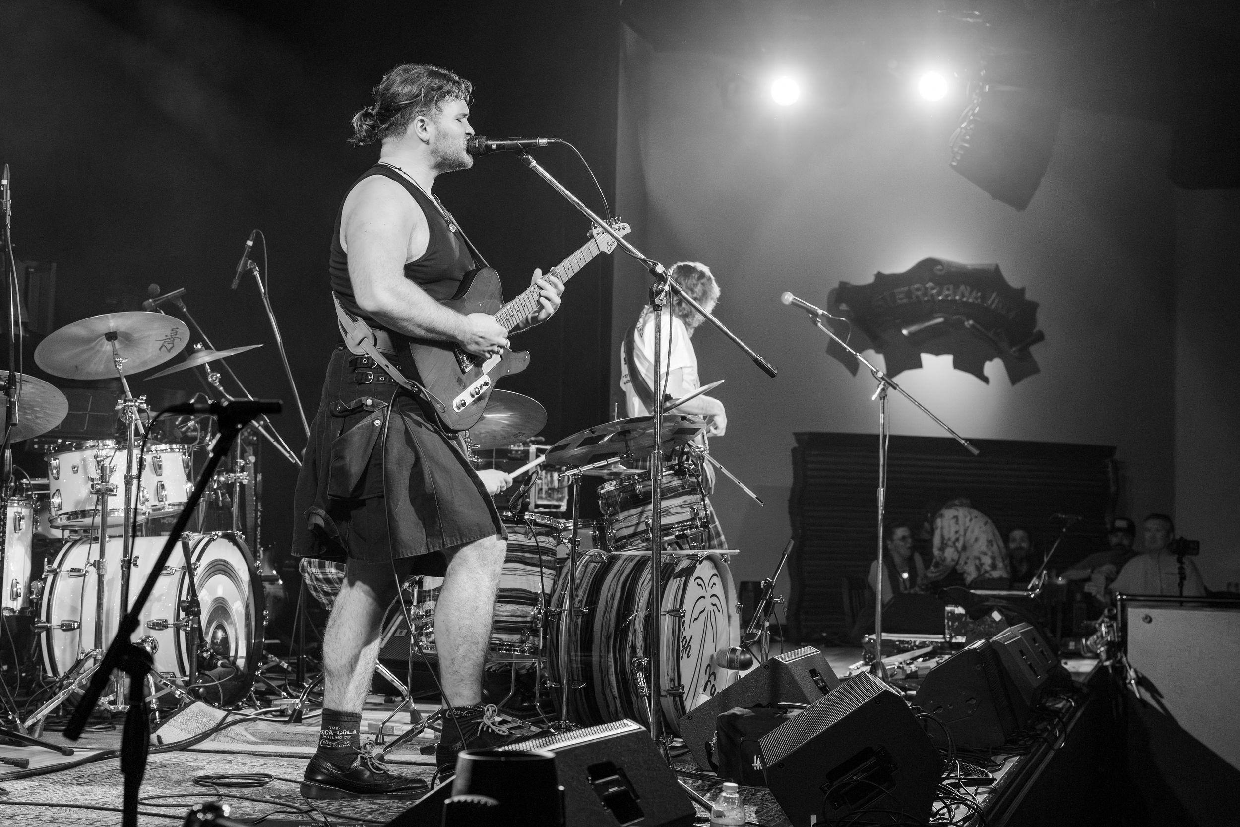 A black and white concert photo of a performer in a kilt playing guitar on stage with drums and equipment visible.