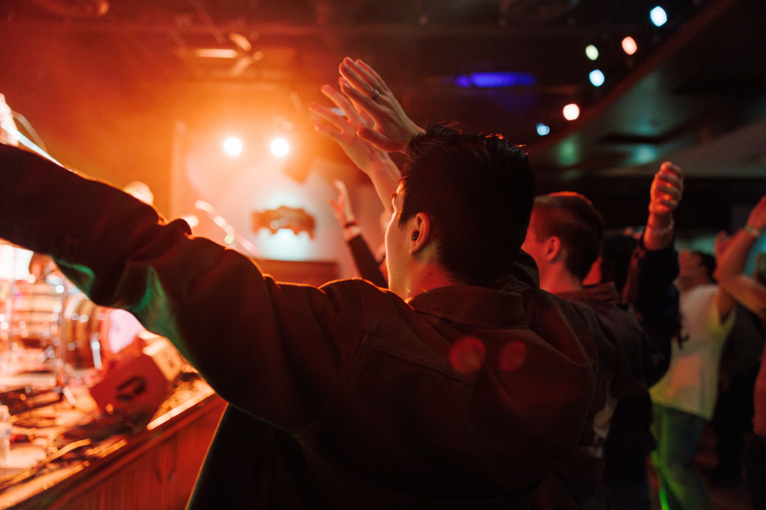 People dancing with their arms raised in a dimly lit nightclub with glowing orange and blue lighting in the background.