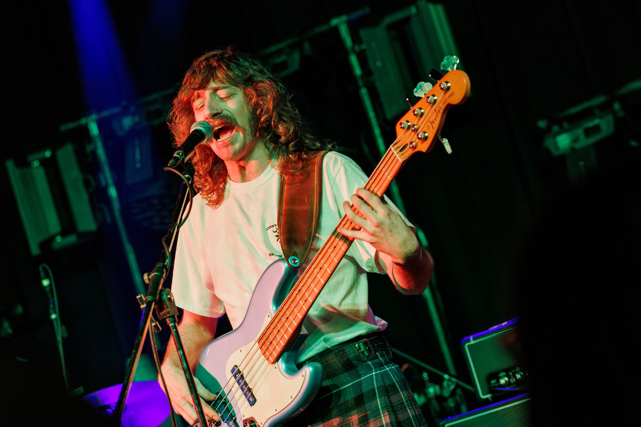 Bass guitarist performs energetically on stage with long curly hair, lit by green and blue stage lighting.