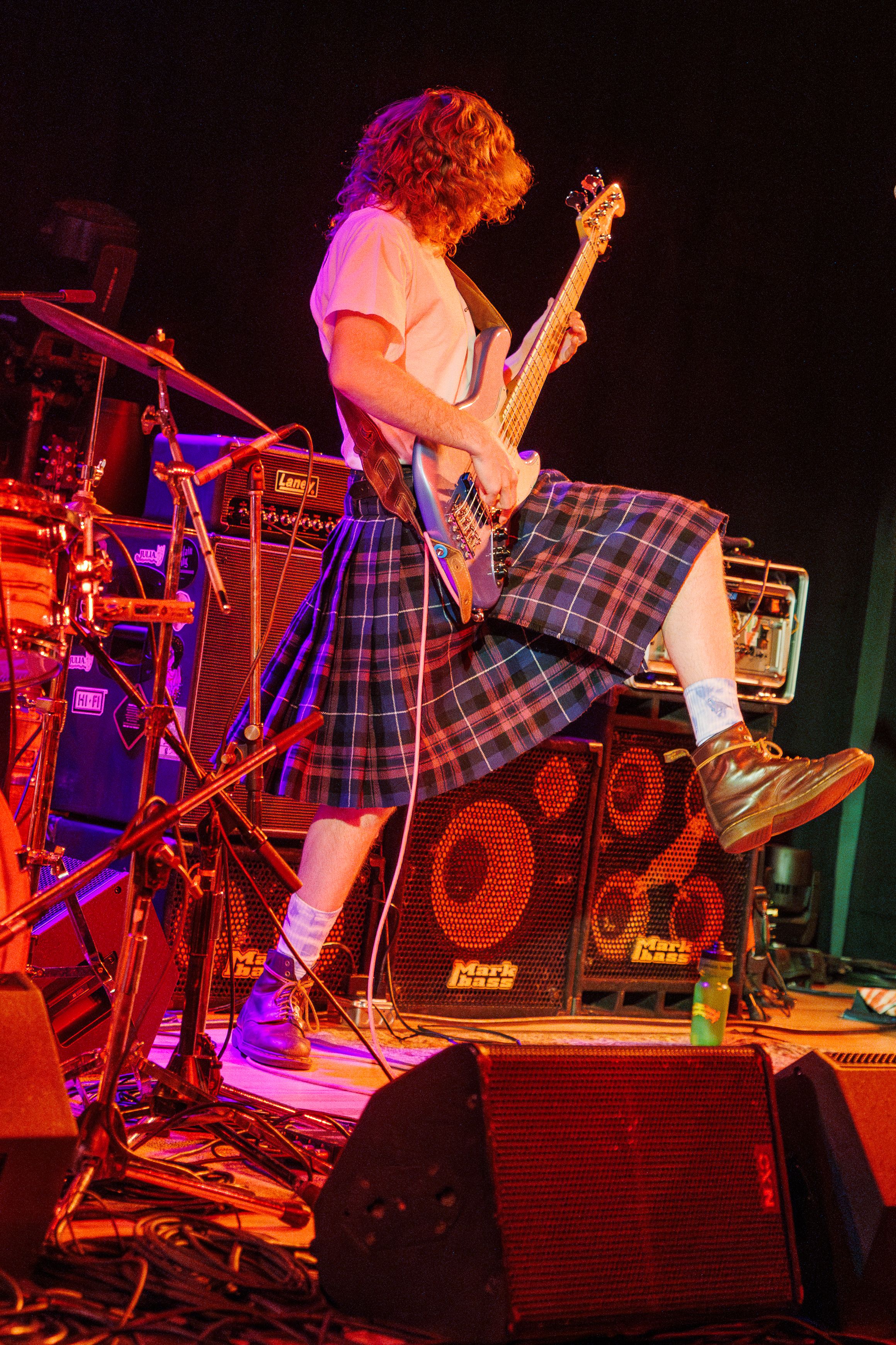 Musician wearing plaid kilt performs energetically on stage with bass guitar under colorful concert lighting.