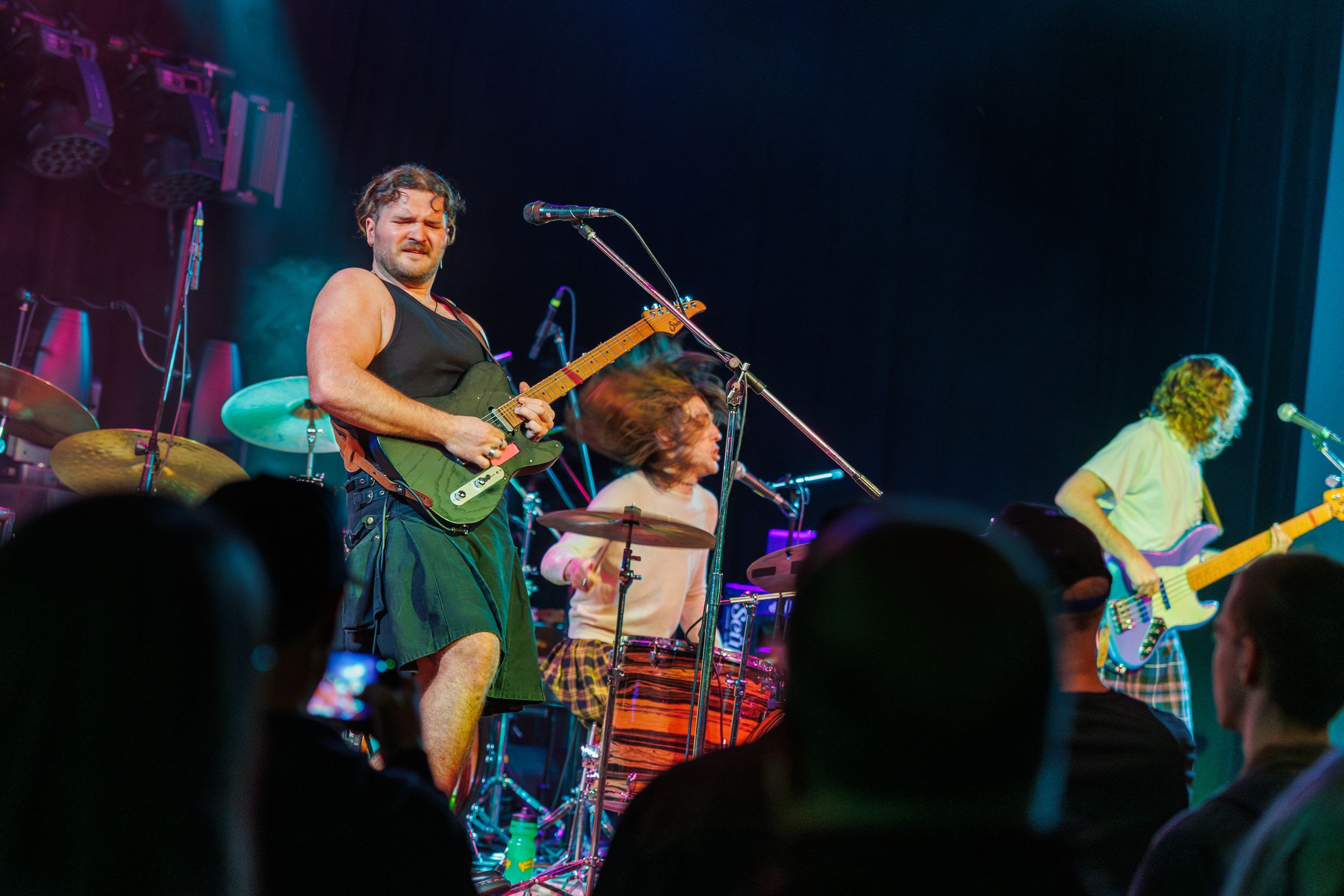 Musicians perform on stage with electric guitars and drums during a live concert with blue stage lighting.