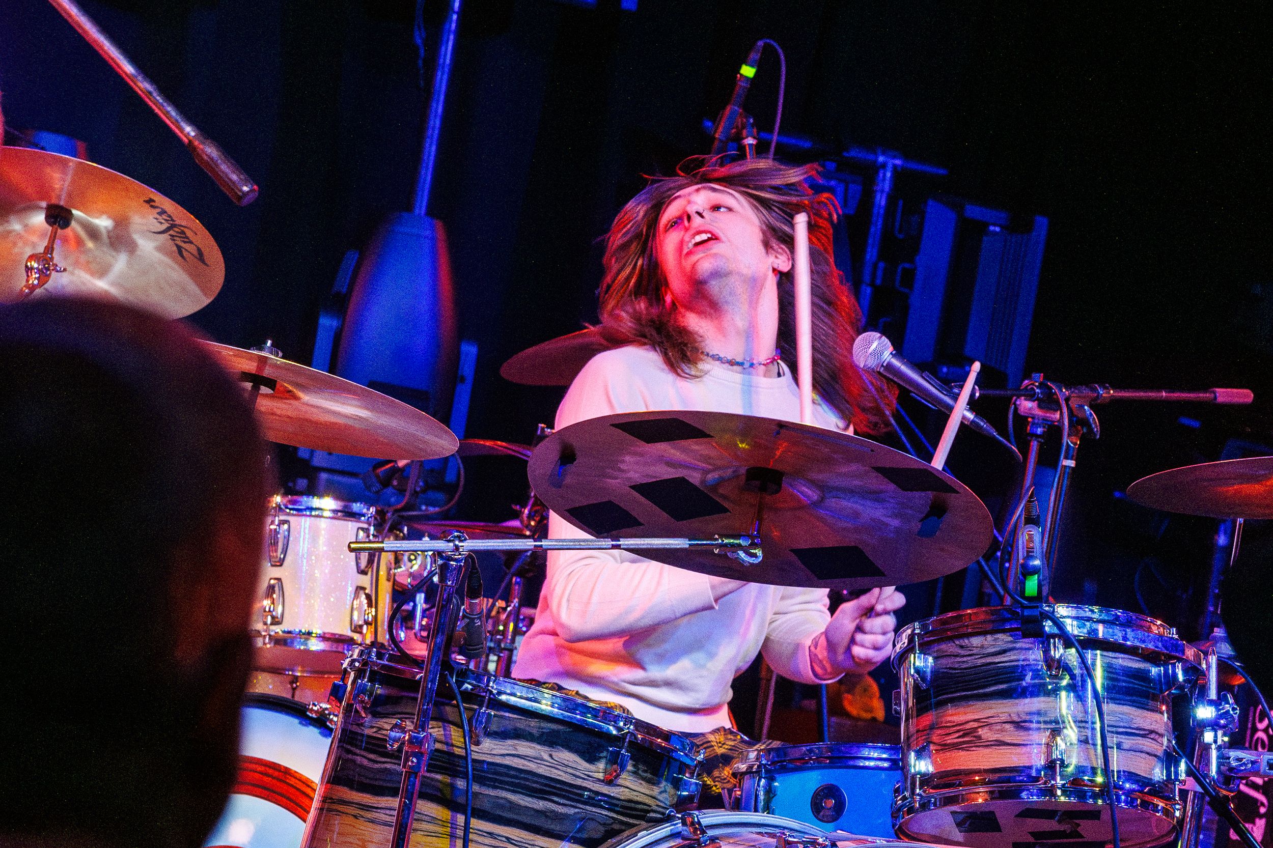 A drummer performs energetically behind a drum kit on stage with dramatic blue concert lighting in the background.